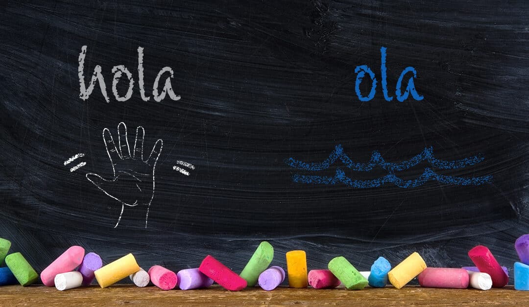 Chalkboard showing Spanish homophones with the word hola and a hand waving next to the word ola with a squiggly wave