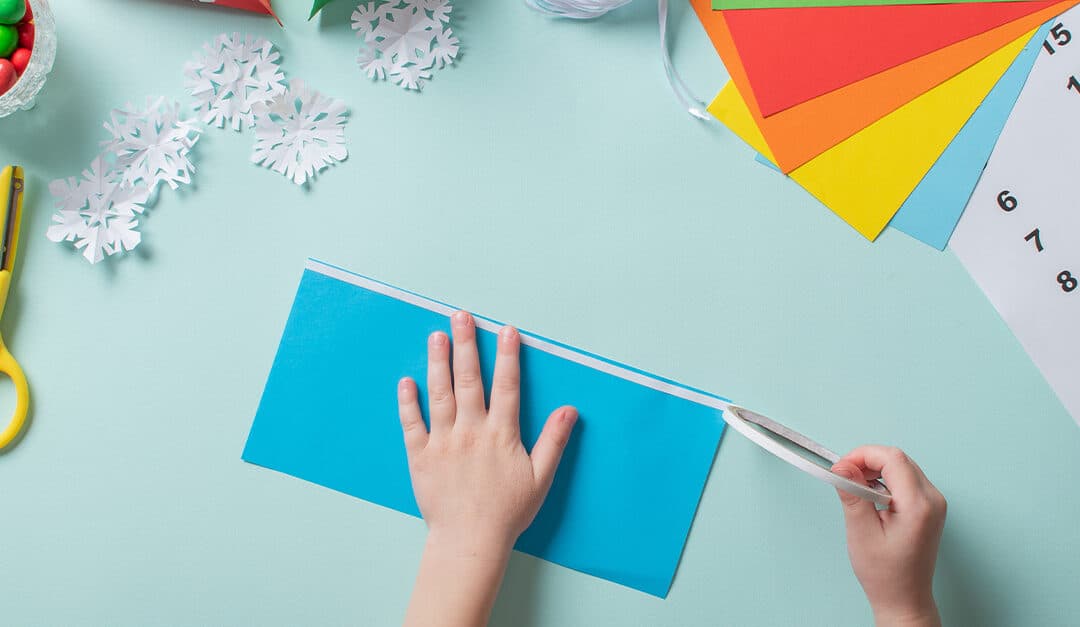 Child's hands creating holiday decorations and calendar with blue and other color papers and white cutout snowflakes - Names in Spanish of Different Holidays