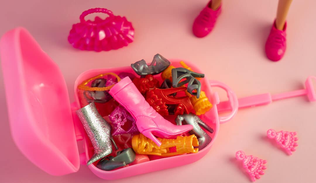 Barbie doll legs in beautiful heels and pink bag open with accessories spilling out on light pink background, closeup