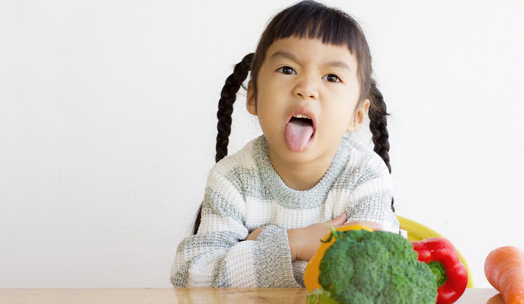 Young girl with expression of disgust, sticking out her tongue while sitting at a table with broccoli, carrots, and bell peppers