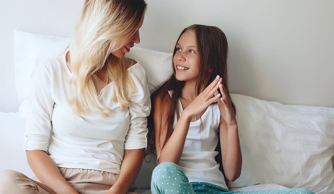 Mom and young daughter sitting next to each other on a bed, talking and smiling