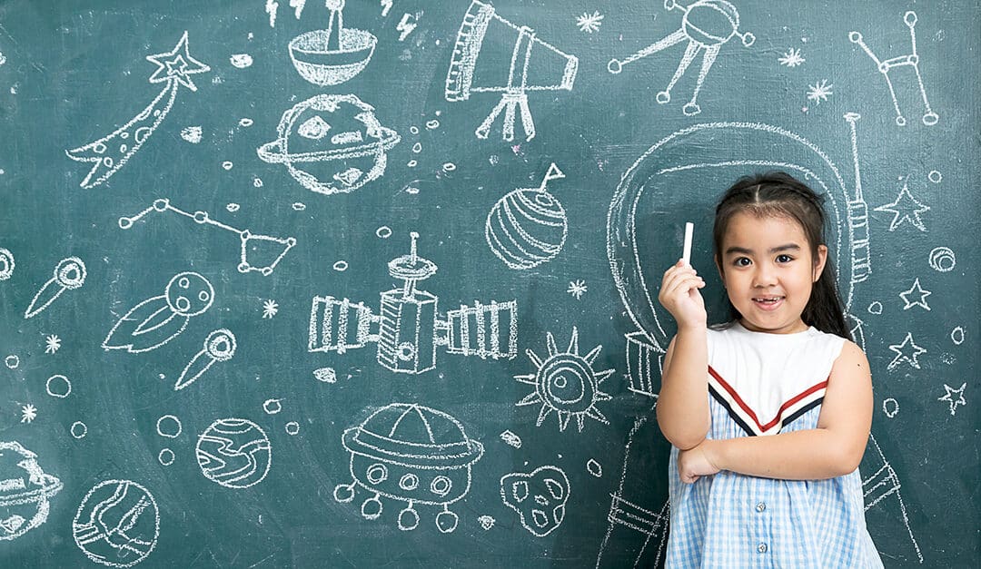 Little girl holding chalk in front of a chalkboard with an outline of an astronaut around her and drawings of space related items scattered across the board, illustrating the blog topic of How to Use the Spanish Phrase "Cuando Sea Grande"