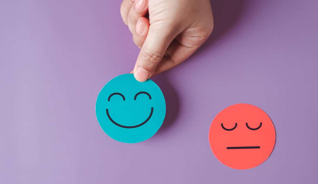 Hand holding cutout of a blue smiley face against a purple background with a red cutout of a face with a neutral expression next to it, illustrating the blog topic Spanish for Emotions