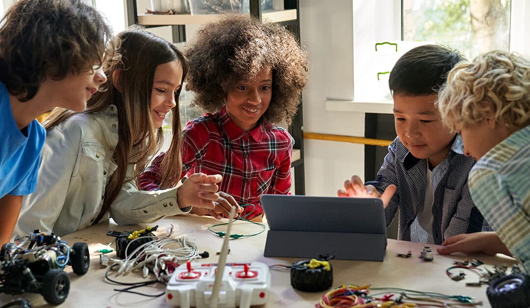 Happy diverse school children building robotic vehicle using tablet. Kids learning programming sitting at table at STEM coding engineering class. Illustrating the blog topic Spanish STEM Vocabulary and Verbs