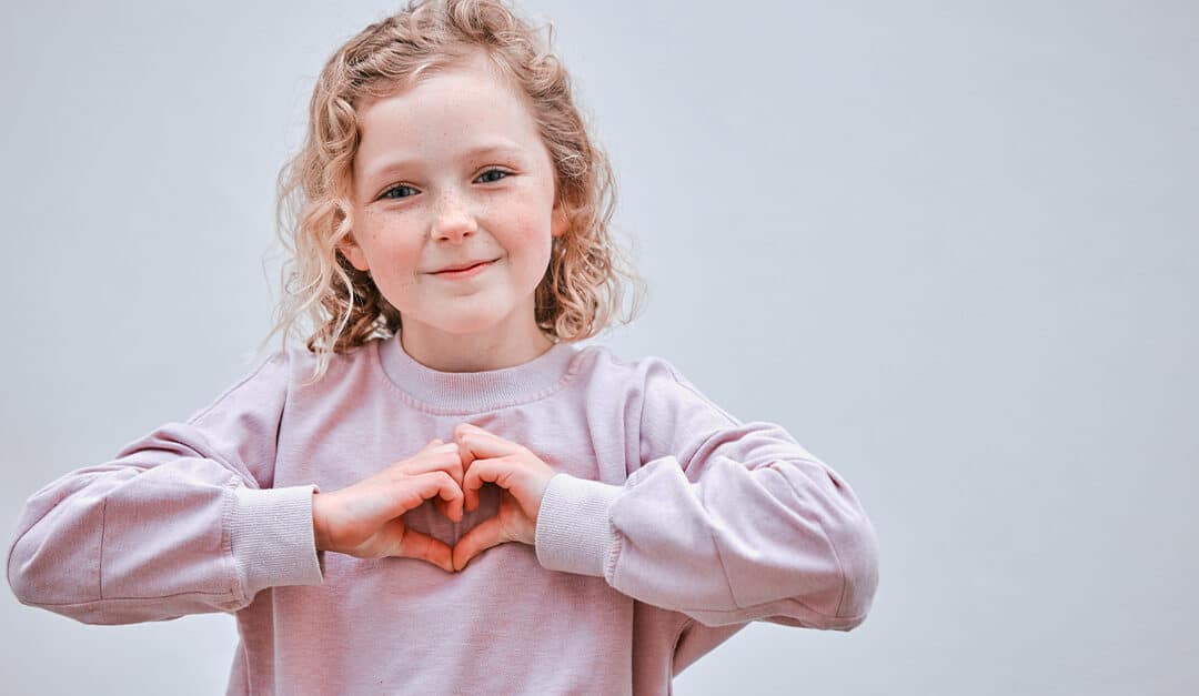 Young girl against a neutral solid background, smiling and making a heart shape with both hands, illustrating the blog topic "Teaching Kids “Please” and “Thank You” in Spanish: Por Favor and Gracias"