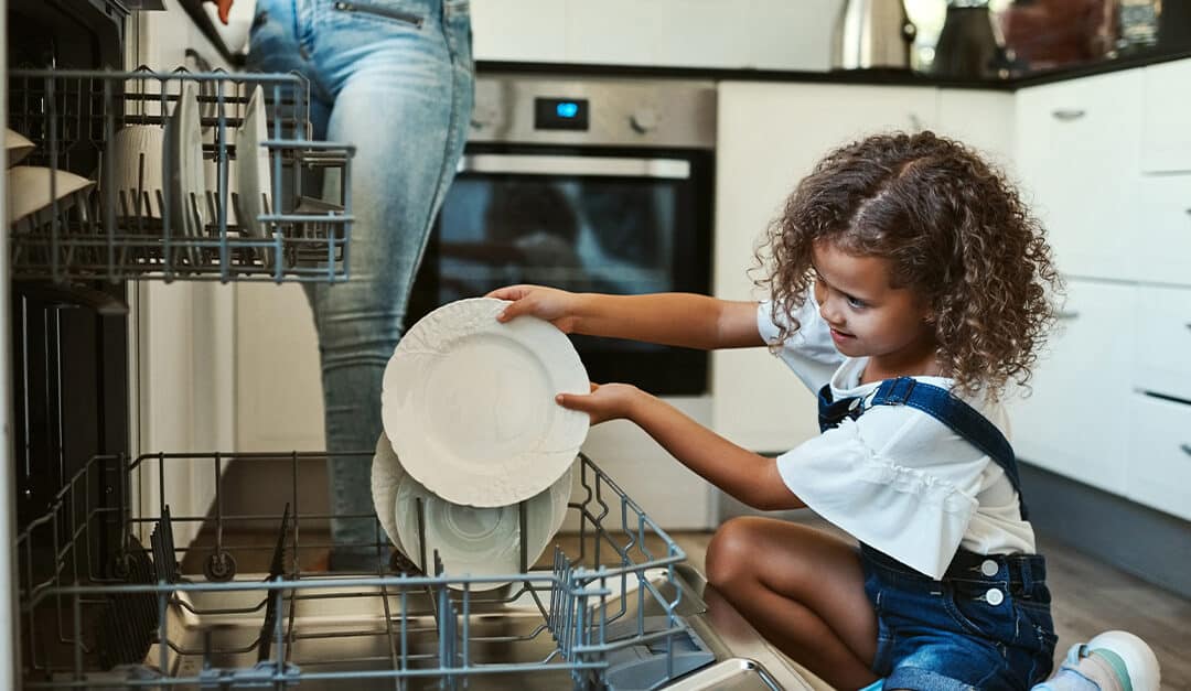 Smiling young girl helping with chores by loading a plate into a dishwasher with her mother in the background