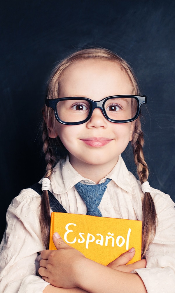 Portrait of a young girl with pigtails and glasses in front of a chalkboard hugging a yellow book entitled "Español"