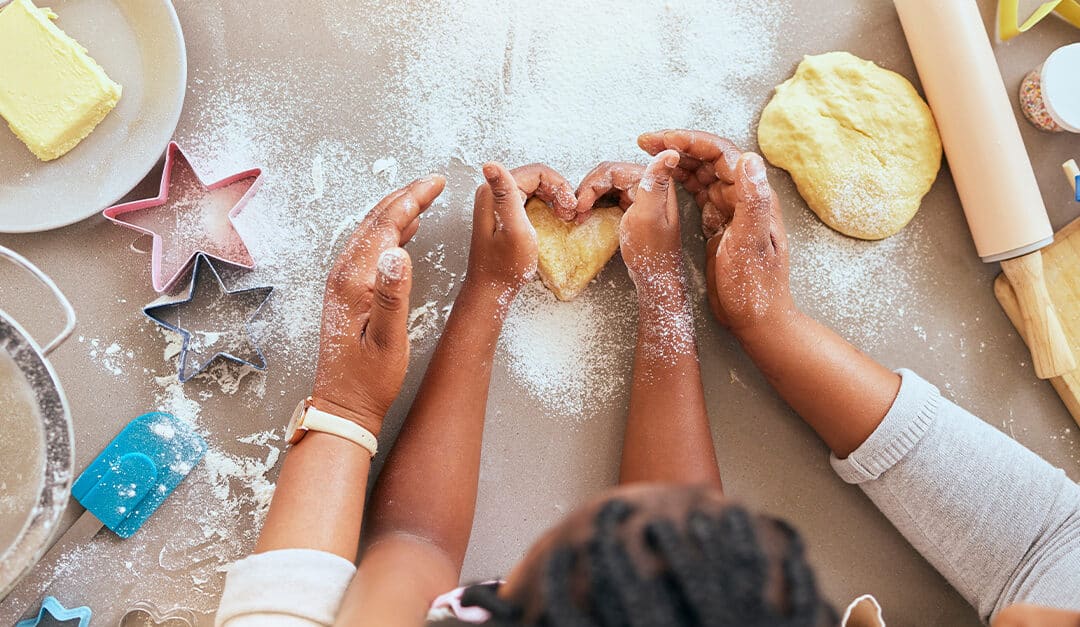 Top view of a mother and daughter making cookies together and shaping dough in the shape of a heart with a rolling pin, butter, flour, cookie cutters and spatulas