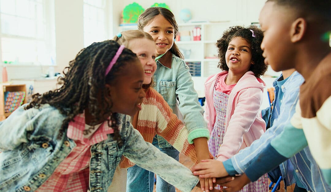 Group of children in an elementary school classroom huddled with hands stacked, smiling and talking
