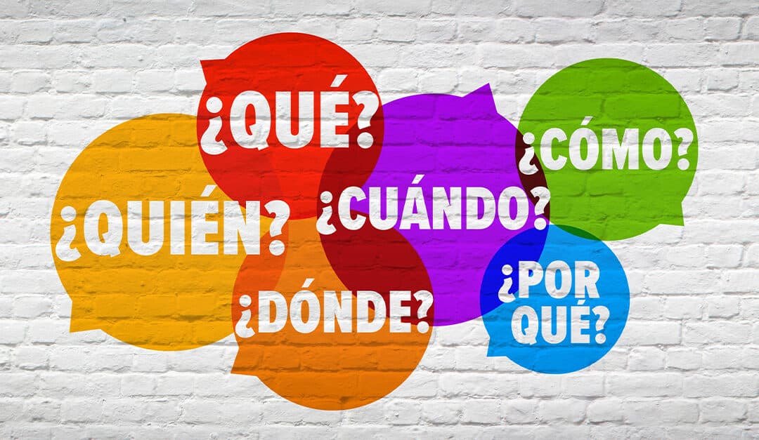 White brick background with different brightly colored speech bubbles containing basic Spanish words like cómo, qué, cuándo, quién, por qué, and dónde